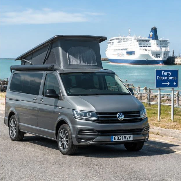 AI-generated illustration: Grey Volkswagen Transporter T6 camper van beside coastal ferry departure area and white ferry, daylight.